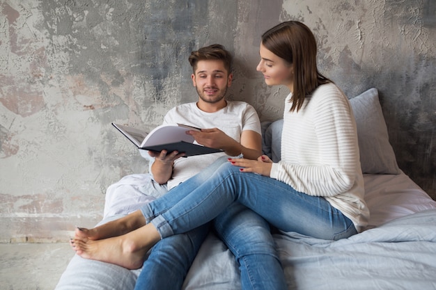 Jong gelukkig lachend paar zittend op bed thuis in casual outfit leesboek dragen jeans, man en vrouw romantische tijd samen doorbrengen