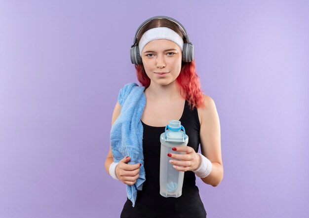 Jong geschiktheidsmeisje in sportkleding met hoofdtelefoons op haar hoofd en met handdoek op haar hals die fles water glimlachend status over purpere muur houden