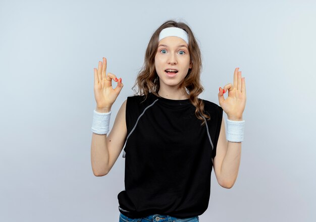 Jong geschiktheidsmeisje in zwarte sportkleding die met hoofdband glimlachend ok teken tonen die zich over witte muur bevinden