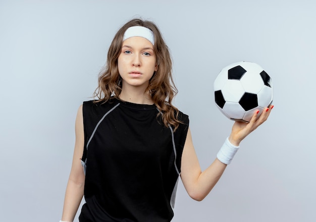 Jong geschiktheidsmeisje in zwarte sportkleding met het voetbalbal van de hoofdbandholding met ernstig gezicht dat zich over witte muur bevindt