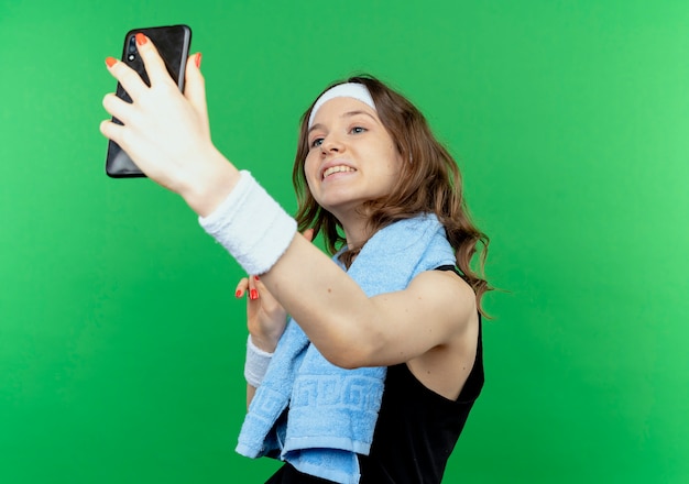 Jong geschiktheidsmeisje in zwarte sportkleding met hoofdband en handdoek om hals die selfie met haar smartphone over groene muur nemen