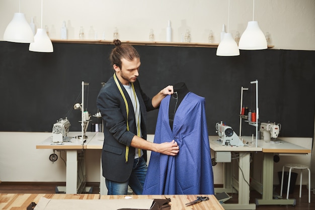 Jong geschoren knappe blanke mannelijke modeontwerper in stijlvolle outfit bezig met nieuwe blauwe jurk voor lentecollectie in zijn atelier. Kunstenaar maken van mooie kleding in zijn atelier