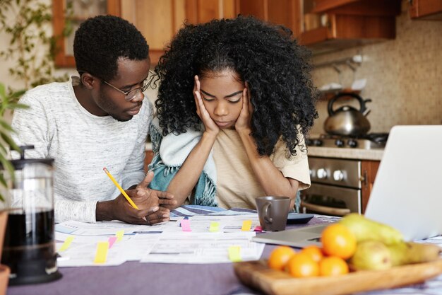 Jong gezin met financieel probleem: gefrustreerde vrouw houdt handen op haar wangen, kijkt wanhopig naar papieren op tafel, kan niet tegen stress, haar man zegt dat alles goed komt