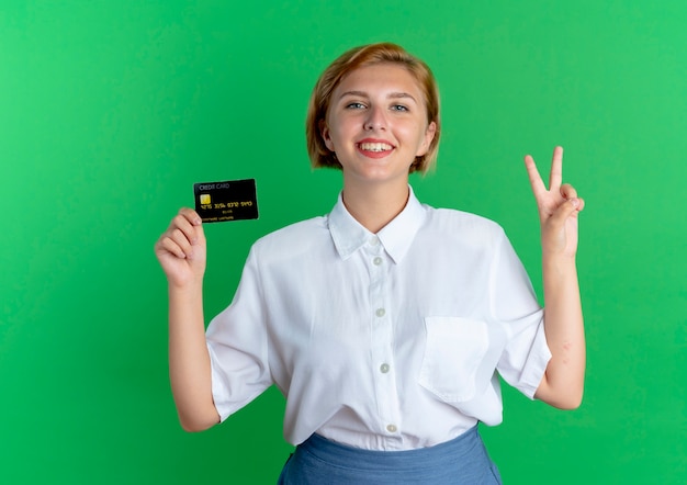 Jong glimlachend blond russisch meisje houdt creditcard en gebaren overwinning handteken