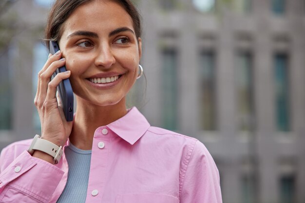 Jong glimlachend vrouwelijk model geniet van internationaal gesprek via smartphonegadget ontvangt oproep in roaminggesprekken over persoonlijke details draagt roze shirt poseert buitenshuis buiten