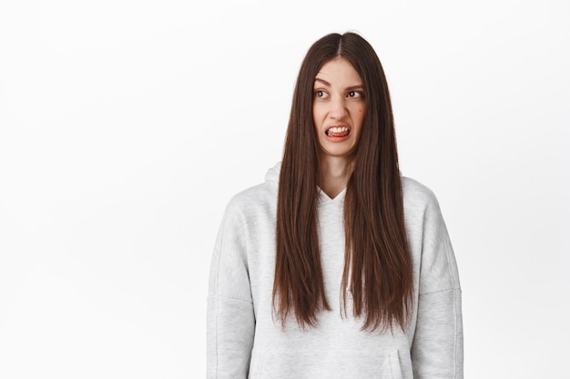 Jong grappig meisje met lang haar, kijk walgend opzij, starend naar ineenkrimpende inhoud, er slecht uitziend vreselijk product met afkeer, tong tonend en grimassend, staande over witte muur