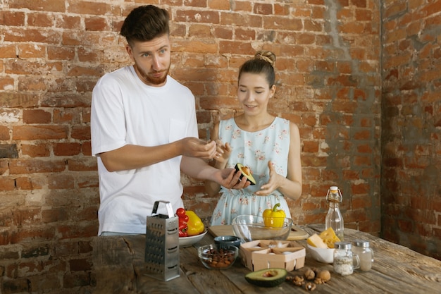 Jong Kaukasisch gelukkig paar die samen koken met behulp van groenten, kaas, eieren en noten in recept tegen bakstenen muur in hun keuken. Voeding, gezonde voeding, gezin, relaties, concept van het huiselijk leven.