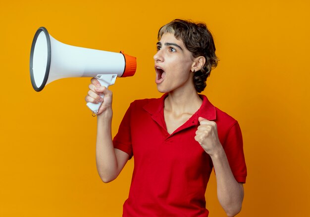 Jong Kaukasisch meisje met pixie kapsel gebalde vuist praten door spreker kijken kant geïsoleerd op een oranje achtergrond