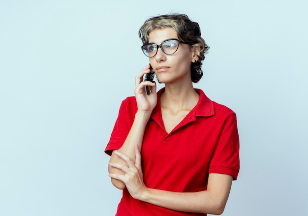 Jong kaukasisch meisje met pixie-kapsel met een bril die aan de telefoon praat en er recht uitziet en de hand op de arm legt