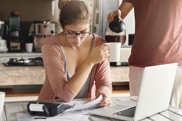 Jong Kaukasisch paar dat financiële problemen heeft. Beklemtoond wijfje die in glazen koffie drinken terwijl het beheren van gezinsbudget, die bij keukentafel zitten met documenten, notitieboekjecomputer en calculator