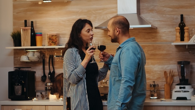 Jong kaukasisch paar flirten tijdens feestelijk diner met tafel op de voorgrond. volwassenen die een romantische date hebben thuis in de keuken, rode wijn drinken, praten, lachend genietend van de maaltijd in de eetkamer