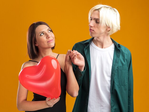 Jong koppel blij vrouw en zelfverzekerde man op Valentijnsdag hand in hand vrouw met hartvormige ballon opzoeken man kijken naar haar geïsoleerd op oranje muur