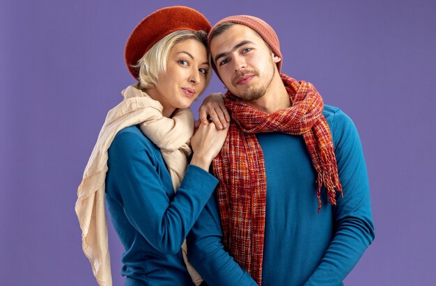 Jong koppel dragen hoed met sjaal op Valentijnsdag lachend meisje handen op de schouder van de man geïsoleerd op blauwe achtergrond