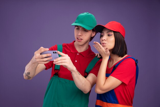 Jong koppel geconcentreerd man serieus meisje in bouwvakker uniform en pet nemen selfie samen meisje houdt hand op de schouder van de man en stuurt klap kus