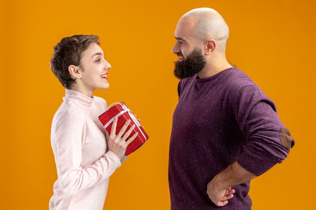 Jong koppel in casual kleding glimlachend bebaarde man een cadeau geven aan zijn verrast en gelukkig vriendin vieren valentijnsdag staande over oranje muur