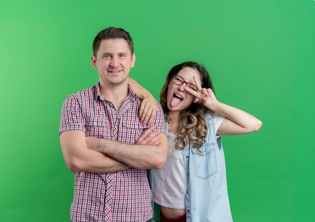 Jong koppel man en vrouw in vrijetijdskleding gelukkige man naast zijn lachende vrolijke vriendin tonen v-teken staande over groene muur