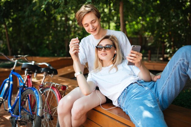 Jong koppel plezier terwijl ze tijd doorbrengen in het park met twee fietsen in de buurt Portret van jongen zittend op een bankje in het park gelukkig kijken naar meisje met blond haar leunend op hem met mobiele telefoon in de hand