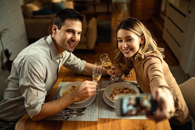 Jong koppel plezier tijdens het roosteren met champagne en het nemen van selfie met mobiele telefoon tijdens een maaltijd aan de eettafel