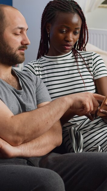 Jong koppel tussen verschillende rassen kijken naar smartphone terwijl zwarte vrouw apparaat thuis vasthoudt. Man en vrouw van gemengd ras die technologie gebruiken en praten terwijl ze op de bank in de woonkamer zitten