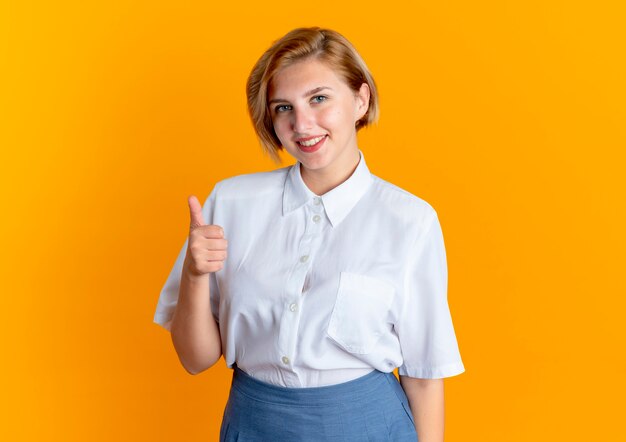 Jong lachend blond Russisch meisje duimen omhoog kijken camera geïsoleerd op een oranje achtergrond met kopie ruimte