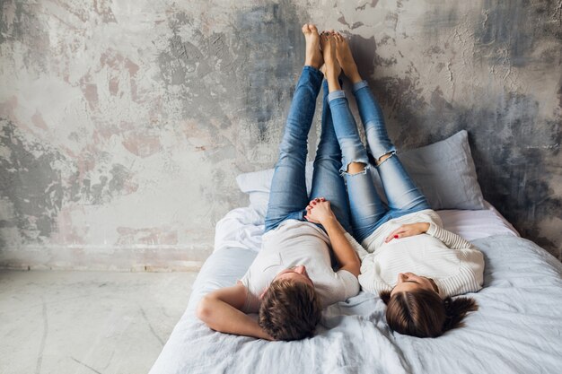 Jong lachend paar liggend op bed thuis in casual outfit, gekleed in spijkerbroek, man en vrouw gelukkige tijd samen doorbrengen, ontspannen, liefde en romantiek