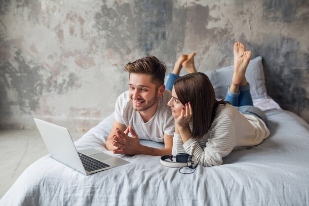 Jong lachend paar liggend op bed thuis in casual outfit, kijken in laptop, man en vrouw gelukkige tijd samen doorbrengen, ontspannen
