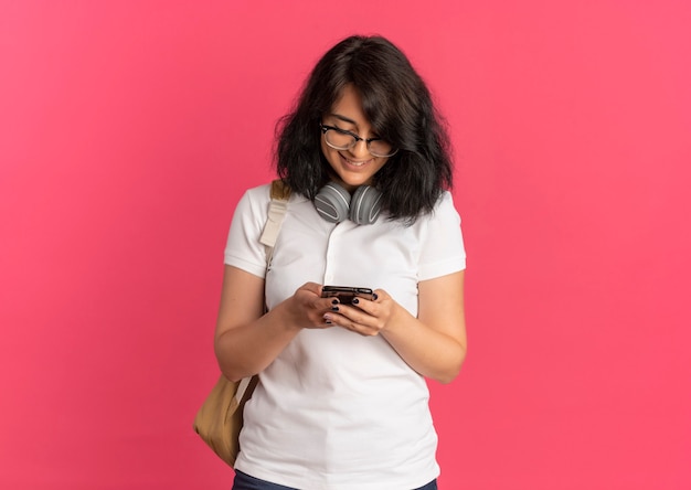 Jong lachend vrij Kaukasisch schoolmeisje bril terug zak en koptelefoon kijkt naar telefoon op roze met kopie ruimte