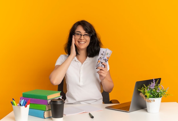 Jong lachend vrij Kaukasisch schoolmeisje met bril zit aan bureau met schoolgereedschap legt hand op gezicht houdt geld geïsoleerd op oranje ruimte met kopie ruimte