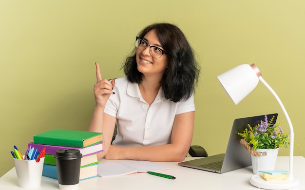 Jong lachend vrij Kaukasisch schoolmeisje met bril zit aan bureau met schoolhulpmiddelen wijst naar kant geïsoleerd op groene ruimte met kopie ruimte