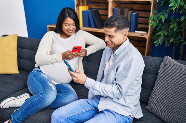 Jong latijns paar verwacht baby met behulp van smartphone zittend op de bank thuis