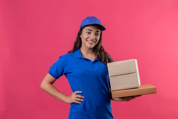 Jong levering meisje in blauw uniform en pet met kartonnen dozen glimlachend zelfverzekerd positief en gelukkig staande over roze achtergrond