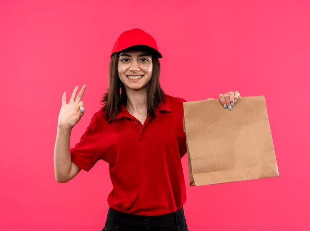 Jong leveringsmeisje die rood poloshirt en GLB dragen die document pakket houden dat met gelukkig gezicht glimlacht dat ok teken toont dat zich over roze muur bevindt