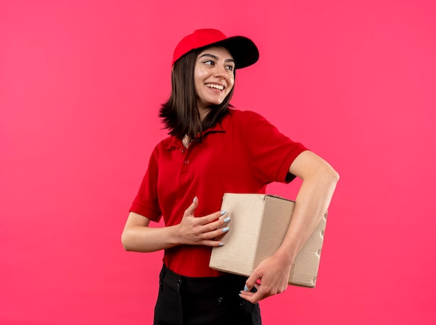 Gratis foto jong leveringsmeisje die rood poloshirt en glb-het pakket van de holdingsdoos dragen die opzij met gelukkige glimlach op gezicht kijken die zich over roze achtergrond bevinden