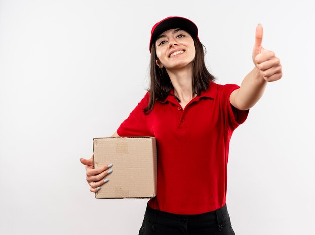 Jong leveringsmeisje die rood uniform en GLB dragen die kartondoos houden die met gelukkig gezicht glimlachen die duimen tonen die zich over witte muur bevinden