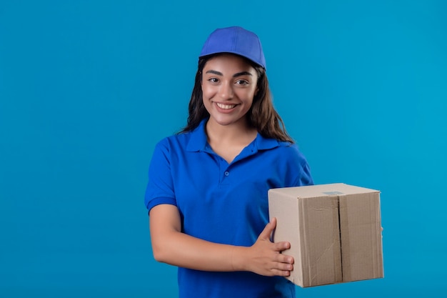 Jong leveringsmeisje in blauw uniform en de doospakket van de GLBholding die camera glimlachen die vriendschappelijk status over blauwe achtergrond bekijken