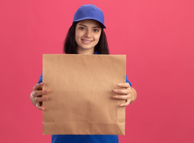 Jong leveringsmeisje in blauw uniform en GLB die document pakket glimlachend zelfverzekerd over roze muur houden