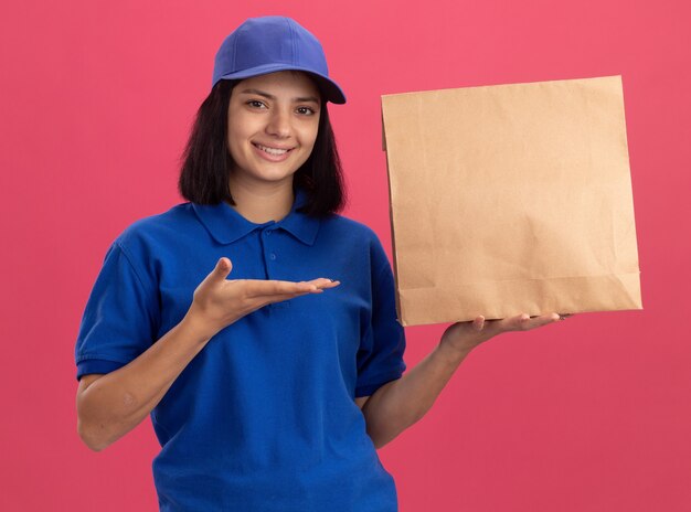 Jong leveringsmeisje in blauw uniform en GLB die document pakket tonen die met wapen glimlachen die zelfverzekerd over roze muur staan glimlachen