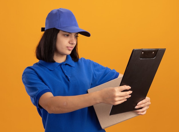 Jong leveringsmeisje in blauw uniform en GLB die kartondoos en klembord houden die het met ernstig gezicht bekijken dat zich over oranje muur bevindt