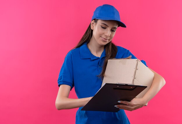 Jong leveringsmeisje in blauw uniform en GLB-doospakket en klembord die blanco pagina's met ernstig gezicht bekijken