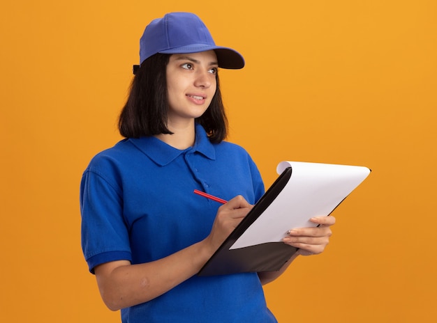 Jong leveringsmeisje in blauw uniform en GLB-holdingsklembord met blanco pagina's en potlood die opzij met glimlach op gezicht kijken dat zich over oranje muur bevindt