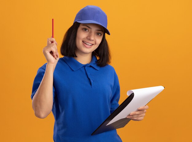 Jong leveringsmeisje in blauw uniform en GLB-holdingsklembord met blanco pagina's en potlood het gelukkige en positieve glimlachen status over oranje muur