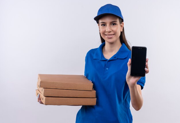 Jong leveringsmeisje in blauw uniform en GLB-holdingsstapel pizzadozen die smartphone zelfverzekerd glimlachen tonen