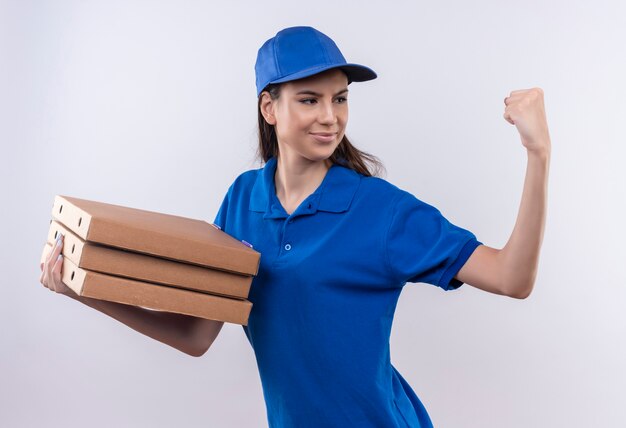 Jong leveringsmeisje in blauw uniform en GLB-holdingsstapel pizzadozen die zelfverzekerd balde vuist, winnaarconcept kijken