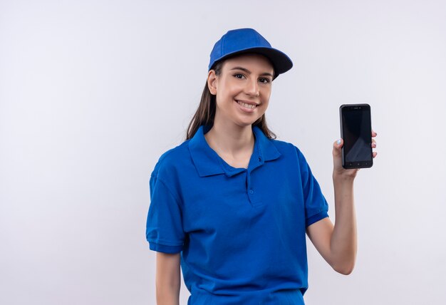 Jong leveringsmeisje in blauw uniform en pet die smartphone tonen die in grote lijnen met gelukkig gezicht glimlacht