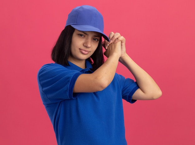 Jong leveringsmeisje in blauw uniform en pet met ernstig gezicht die teamgebaar maken die zich over roze muur bevinden