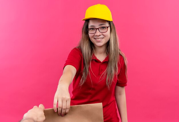 Gratis foto jong leveringsmeisje in rood poloshirt en geel capgiving-document pakket aan een klant vriendelijk glimlachen