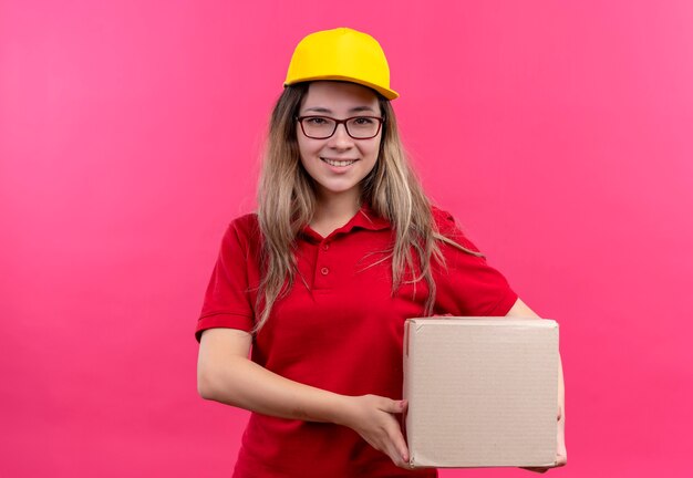 Jong leveringsmeisje in rood poloshirt en geel de doospakket van de glbholding die camera zelfverzekerd glimlachen bekijken