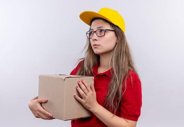 Gratis foto jong leveringsmeisje in rood poloshirt en geel de doospakket van de glbholding die opzij met droevige uitdrukking op gezicht kijken