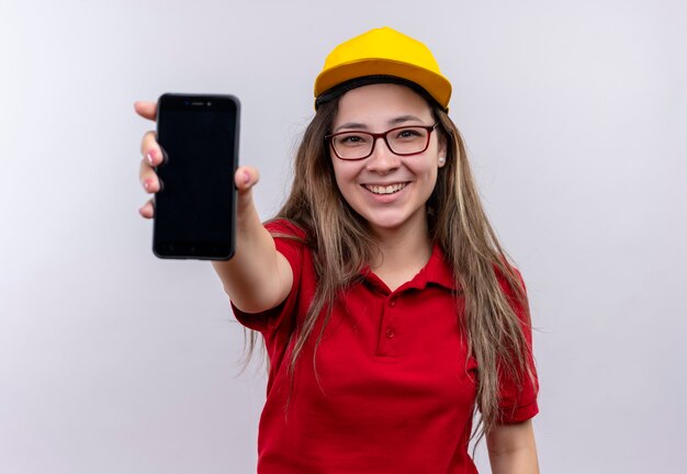 Jong leveringsmeisje in rood poloshirt en geel GLB die smartphone tonen aan camera in grote lijnen glimlachen