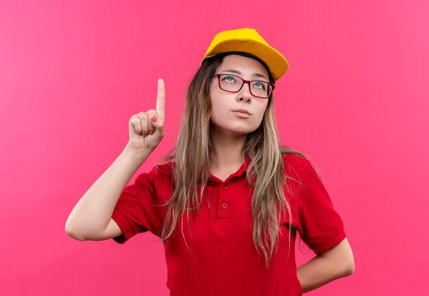 Jong leveringsmeisje in rood poloshirt en geel GLB die wijsvinger tonen die nieuw idee hebben